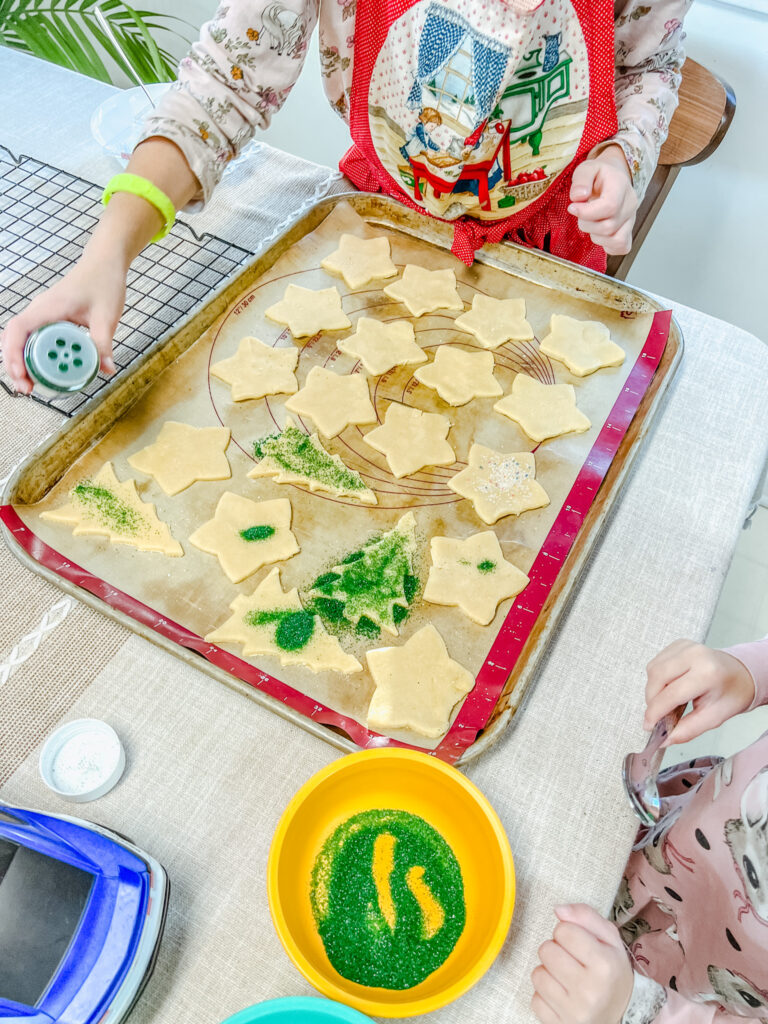 One of my girls sprinkling Sanding Sugar onto the cut sugar cookies on a baking sheet.