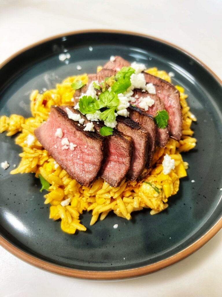 A Picture of medium rare sliced steak on top of the Mexican Street Corn Pasta Salad, topped with queso fresco and cilantro.