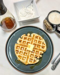 waffles with maple syrup and whipped cream on a blue plate next to a cup of coffee